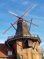 Un molino de viento tradicional con estructura roja y blanca, adosado a un edificio de ladrillo con múltiples ventanas y techo de tejas.
