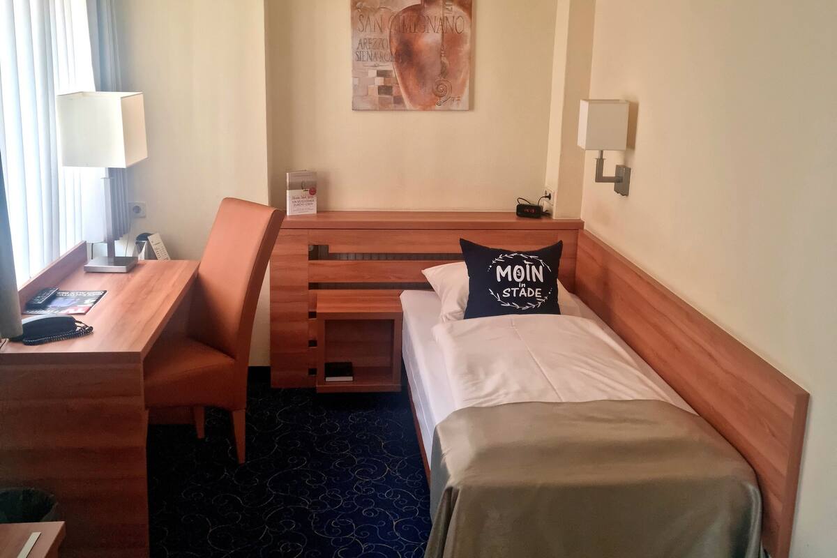 A hotel room with a bed, a desk, a chair, a lamp, a painting on the wall, and a carpeted floor.