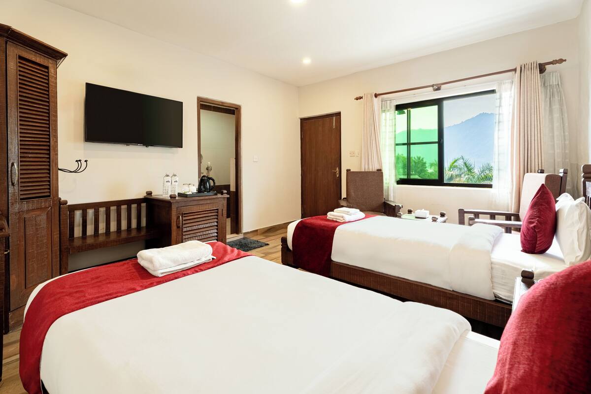 A hotel room with two beds, a TV, a wooden wardrobe, a desk with a kettle, and a window with a mountain view.