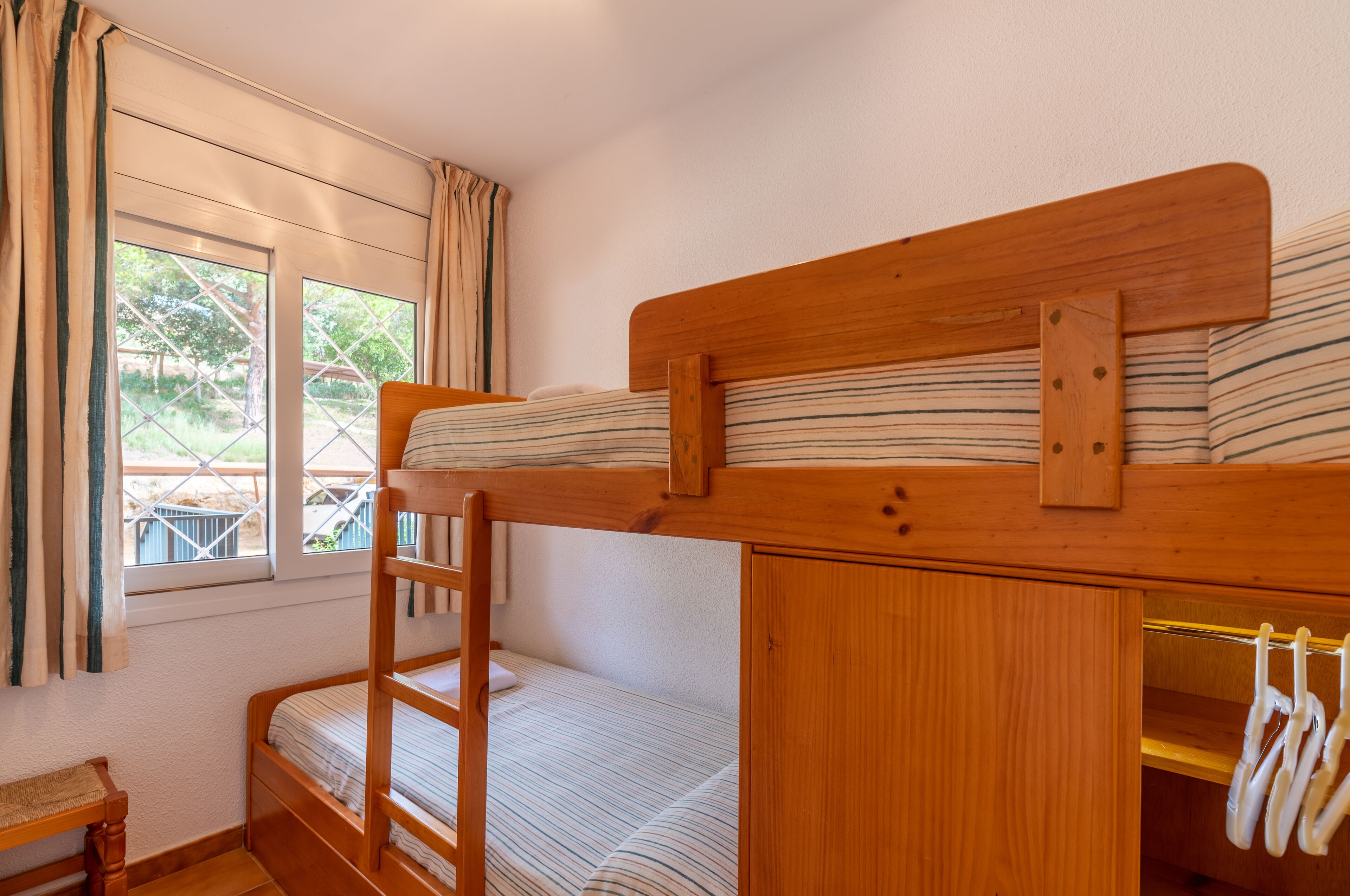 A bunk bed room with a wooden headboard, a small bench, and a window with curtains.