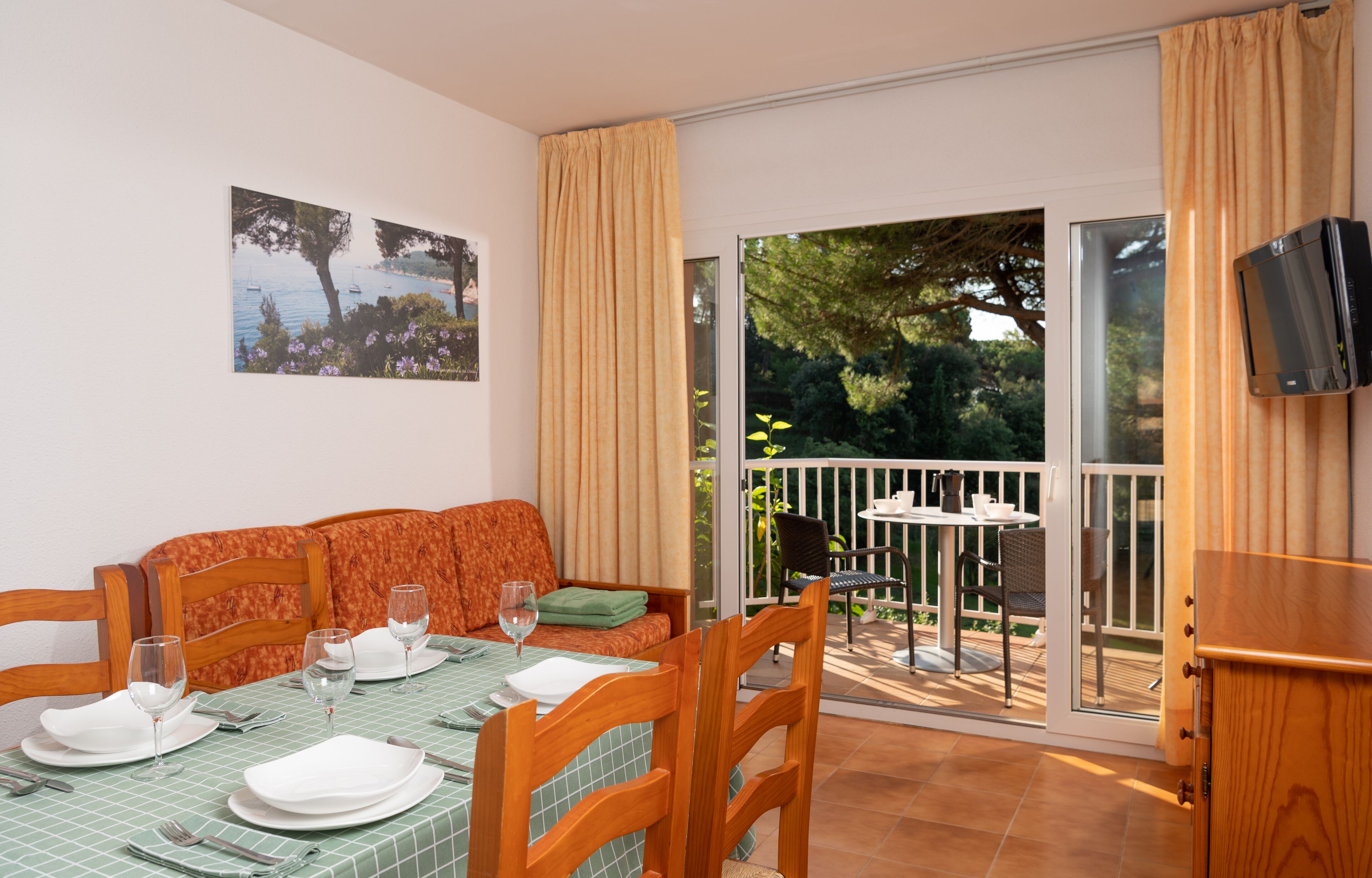 A dining area with a table set for four, chairs, a sofa, and a sliding glass door leading to a balcony with a view of trees and a boat.
