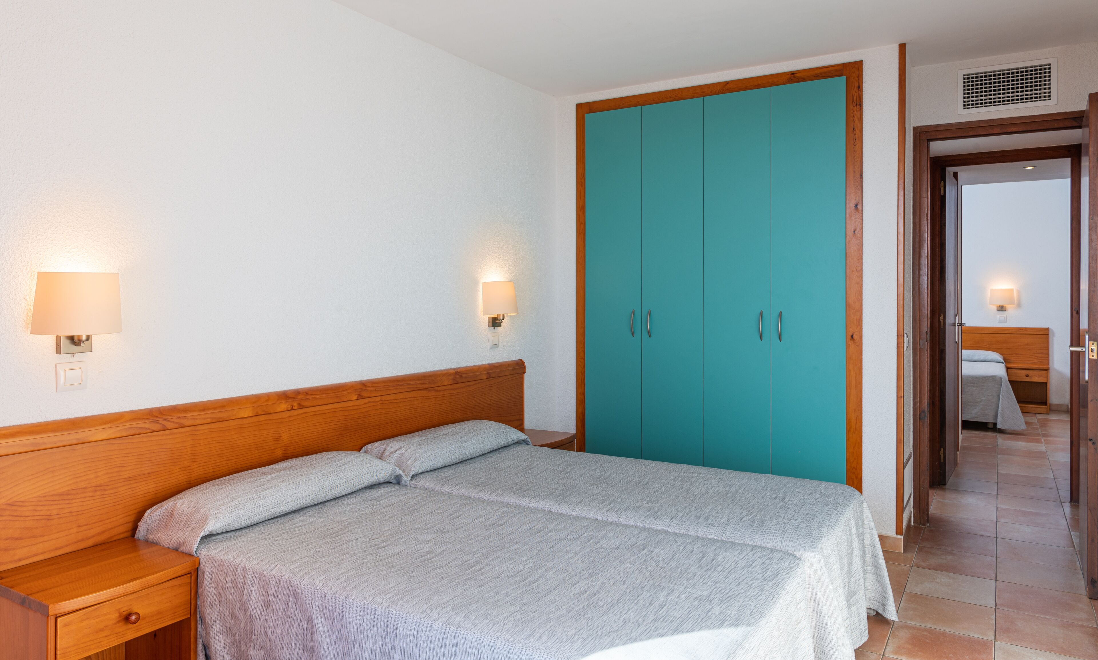 A bedroom with a bed, nightstand, two wall-mounted lamps, and a teal wardrobe.