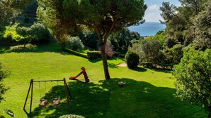 A lush garden with a swing set, a large tree, and a view of the sea. - Albamar Apartaments (Lloret de Mar)