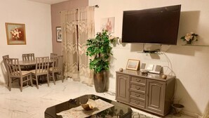 Una sala de estar con televisor, mesa de comedor, sillas, mesa de centro, aparador y una planta en maceta.