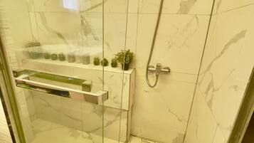Une douche fermée par des parois en verre, dotée d’un pommeau de douche à main, d’une petite étagère avec des plantes et d’un mur recouvert de carreaux de marbre.