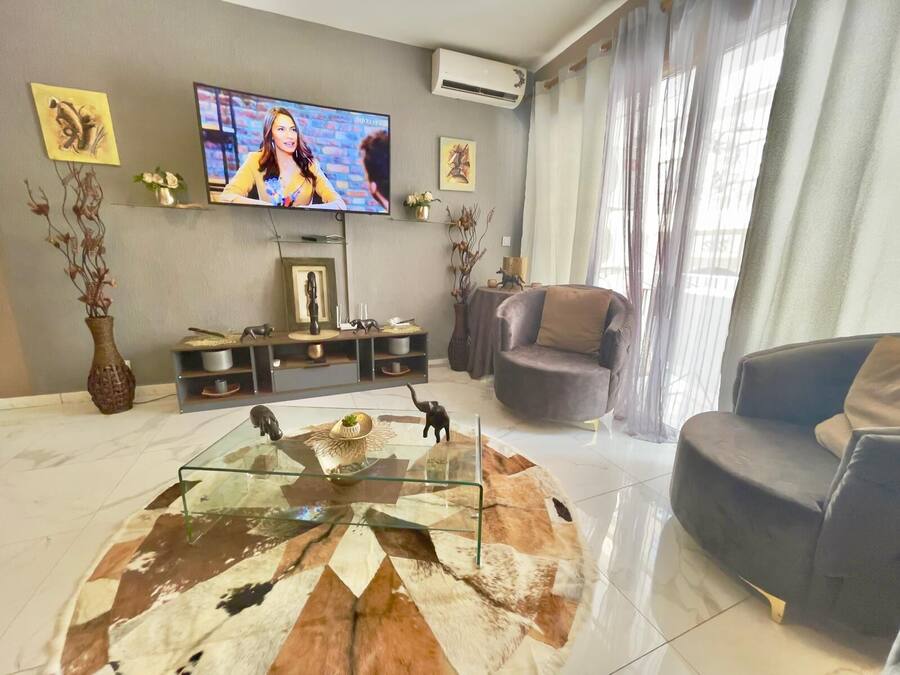 Una sala de estar con un televisor de pantalla plana, una mesa de centro de vidrio con diseño de cuero de vaca, un sofá gris y una consola decorativa con figuritas.