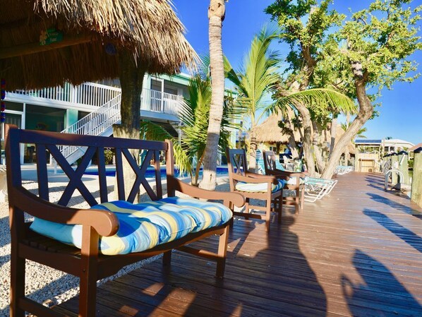 A wooden deck with a bench, palm trees, and a building with a thatched roof.