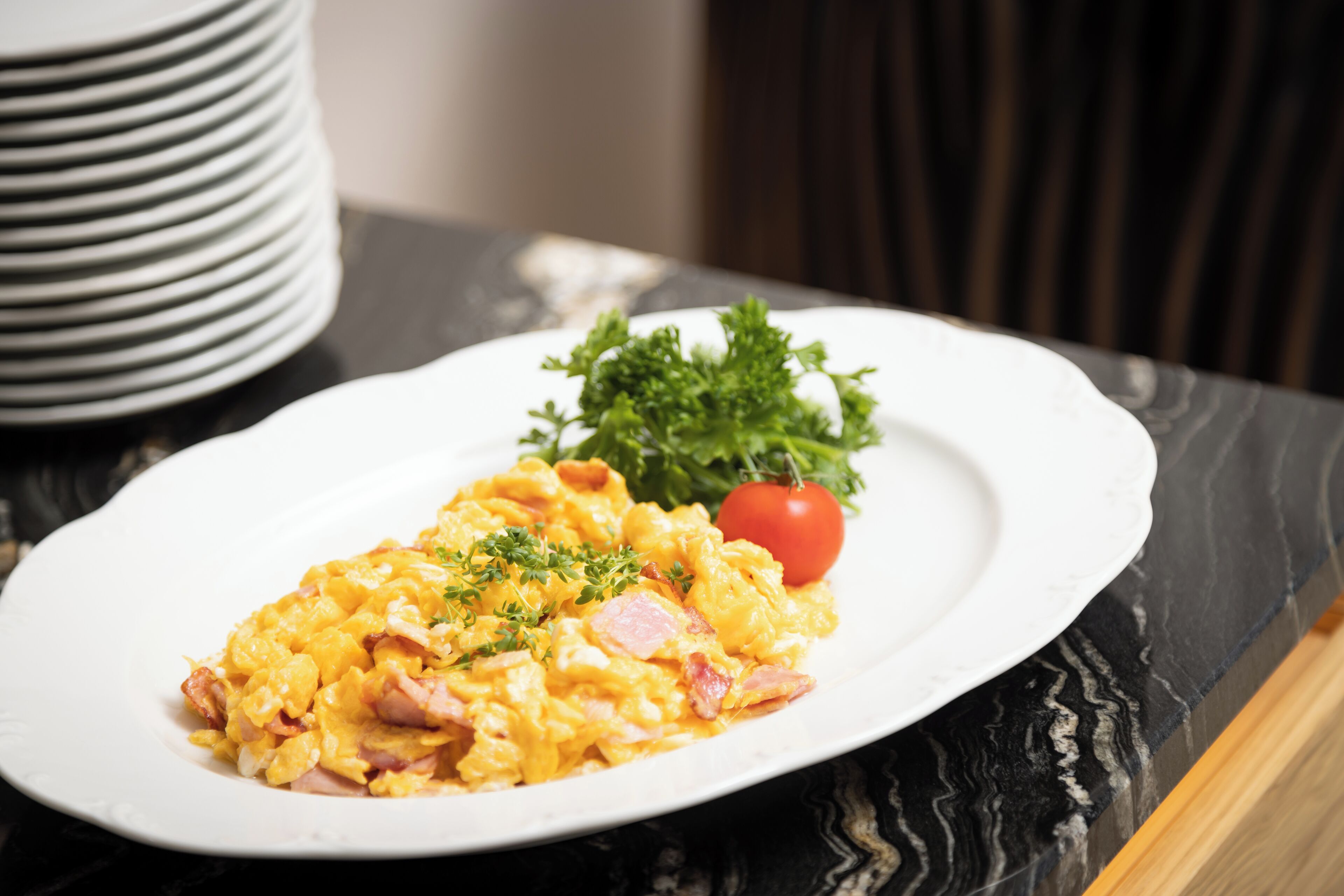 Un plato blanco con huevos revueltos, jamón y una guarnición de perejil, acompañado de un tomate cherry y una ramita de perejil, sobre una encimera de mármol oscuro.