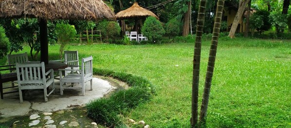 A garden with a thatched-roof gazebo, white wooden chairs, and a small table.