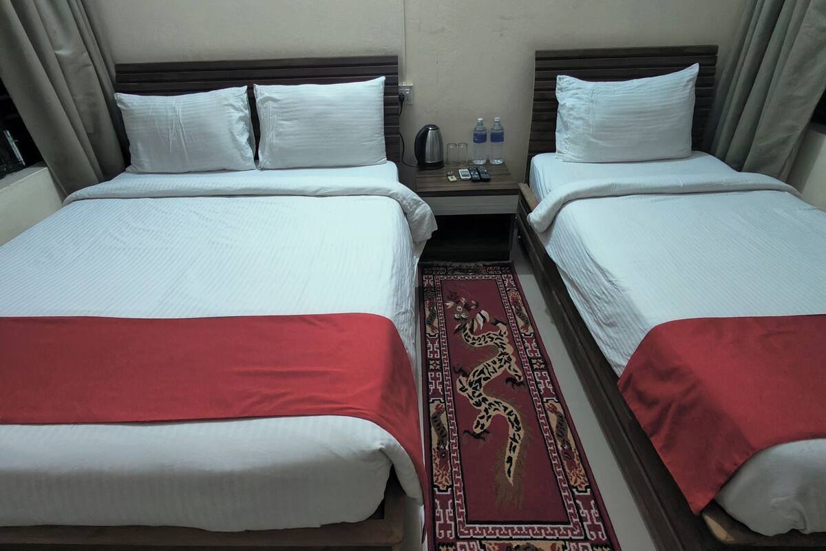 A hotel room with two beds, each with white linens and red bedspreads. There is a small table with a kettle, a remote control, and a glass of water. A decorative rug is placed between the beds.