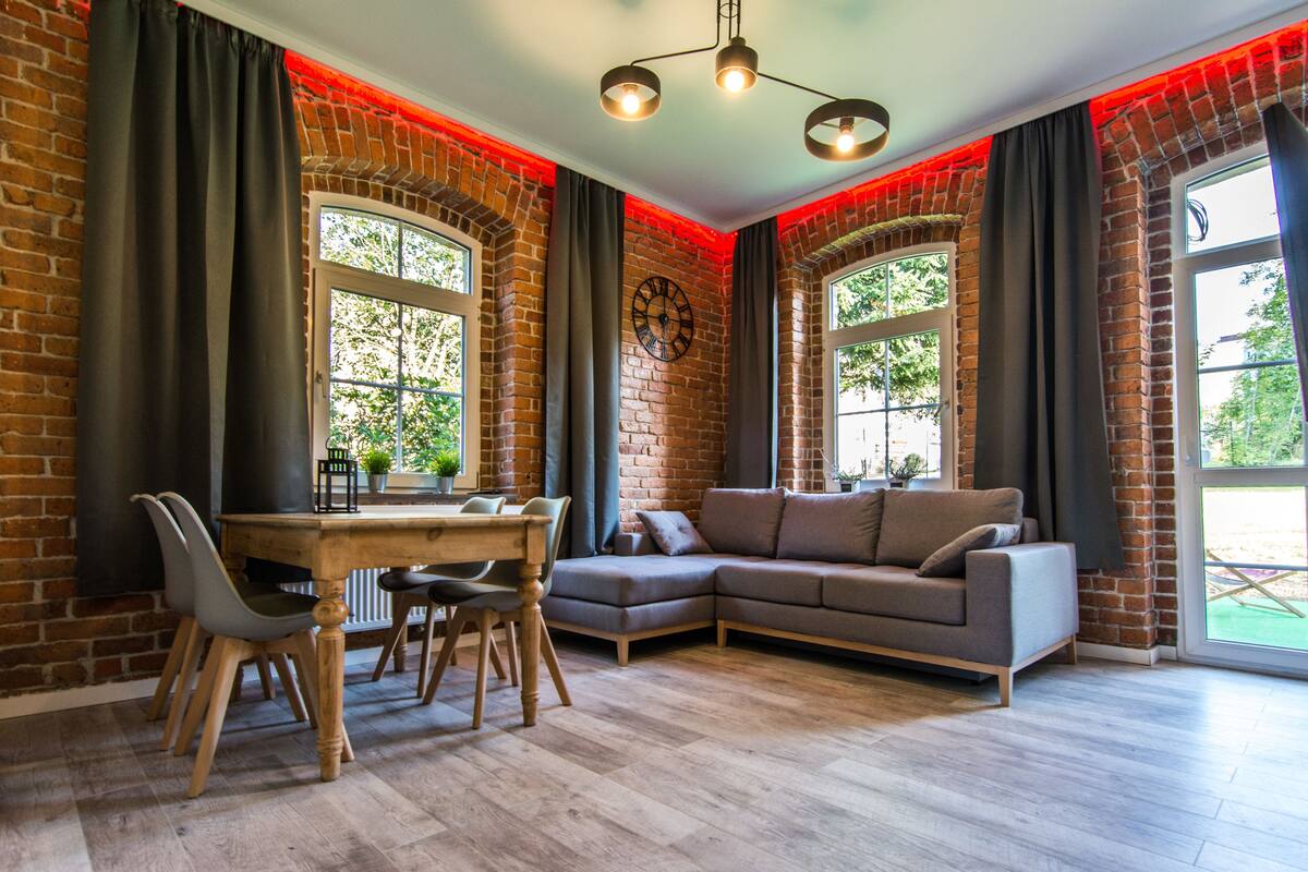 Una sala de estar con pared de ladrillo, una mesa de comedor de madera con sillas, un sofá gris y un ventanal con cortinas.