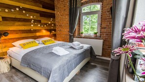 A cozy bedroom with a wooden ceiling, a brick wall, a large window with a view, and a bed with pillows and a blanket.