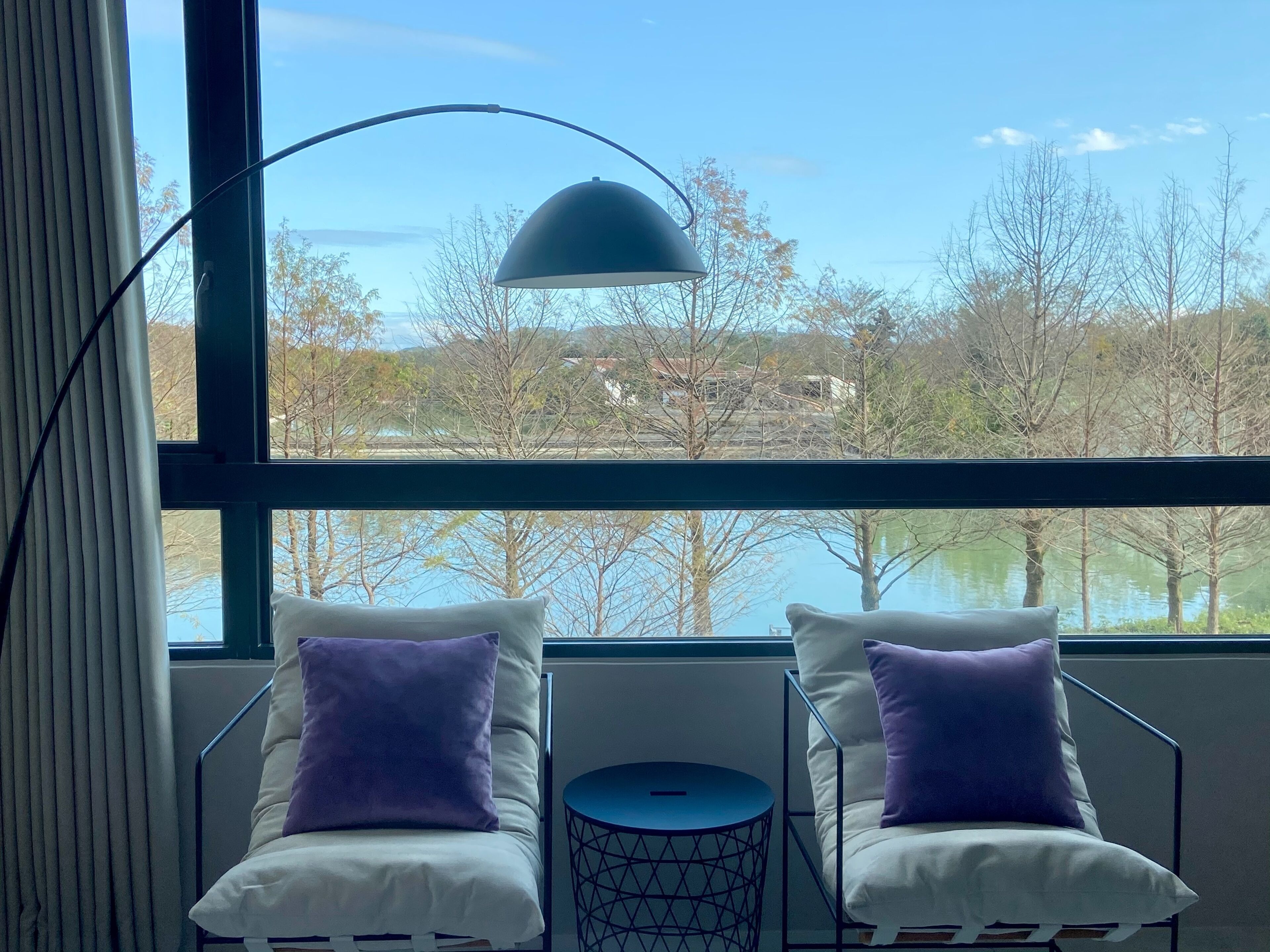A room with two white chairs, each with a purple pillow, and a small round table. A large window provides a view of trees and a body of water.