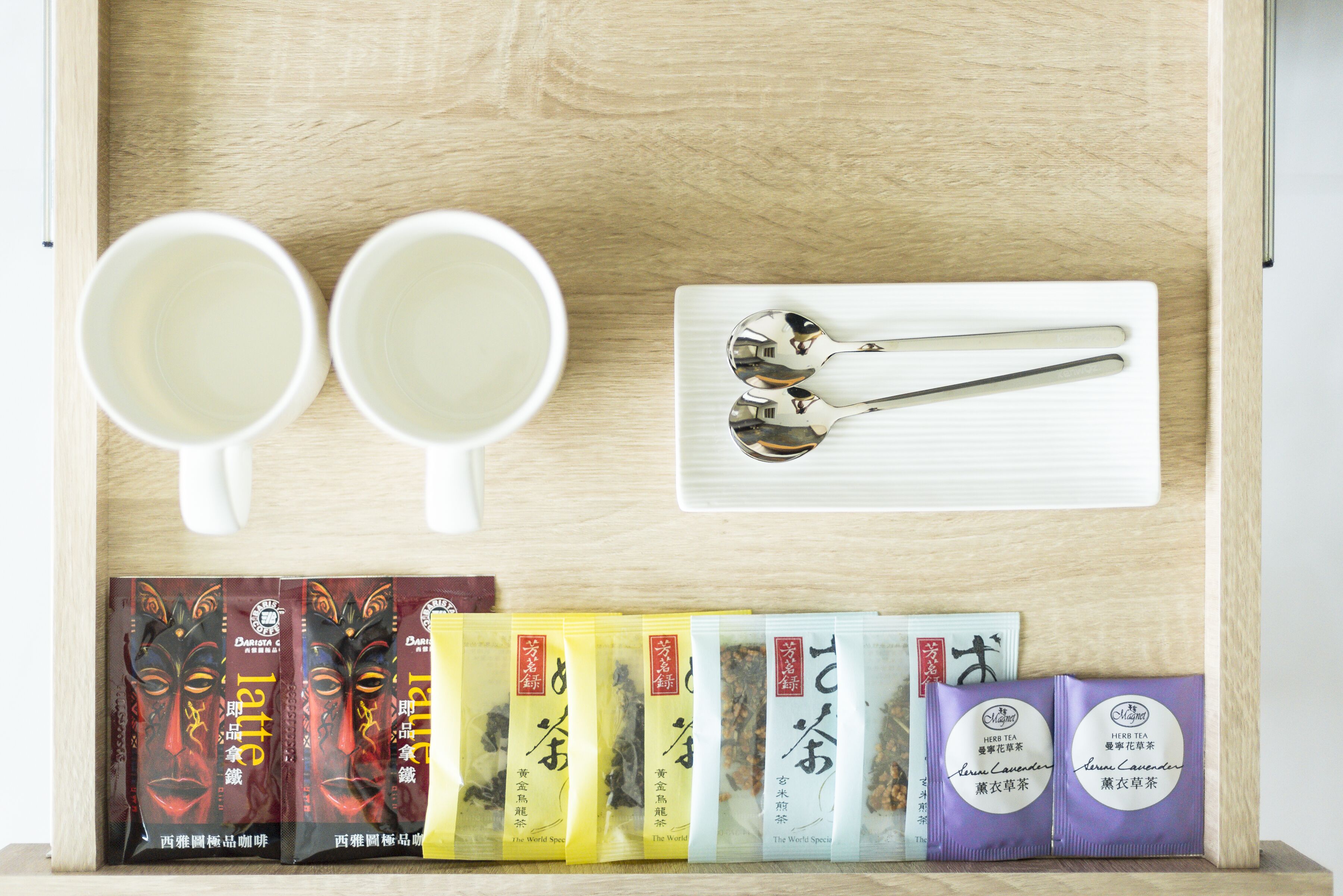 A wooden drawer with two white cups, a pair of spoons, and several tea packets.