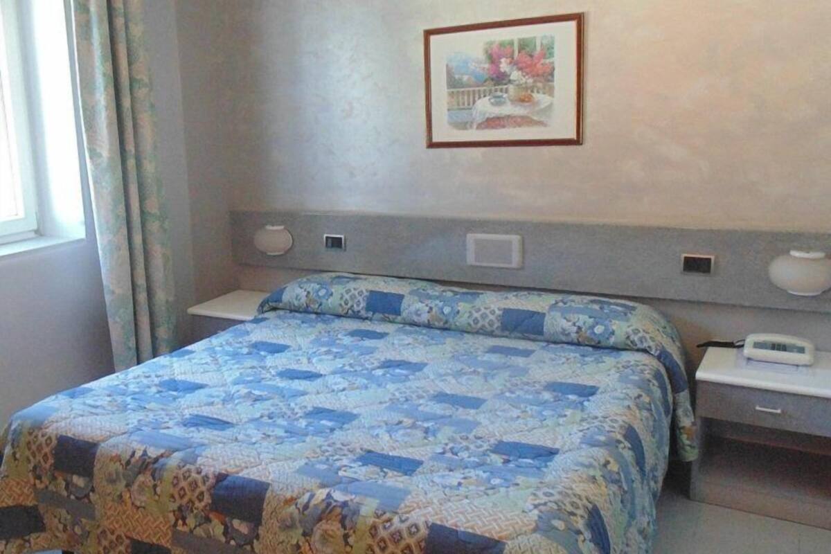 A hotel room with a bed, a bedside table, a nightstand, a framed picture on the wall, and a window with curtains.