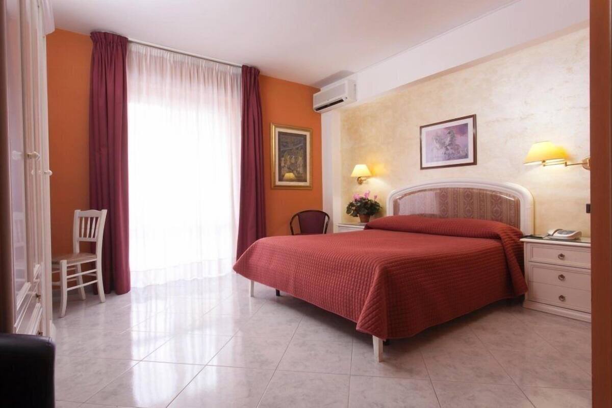 A hotel room with a bed, a chair, a desk with a lamp, and a wall-mounted air conditioner.