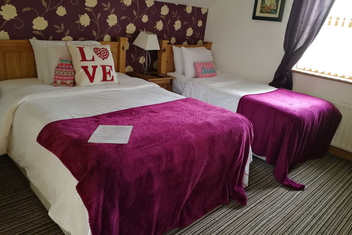 Quarto de hotel com duas camas, cada uma com roupa de cama branca e um cobertor roxo. Almofadas decorativas com a palavra “LOVE” em uma das camas. Uma luminária e um quadro emoldurado estão na mesinha de cabeceira.