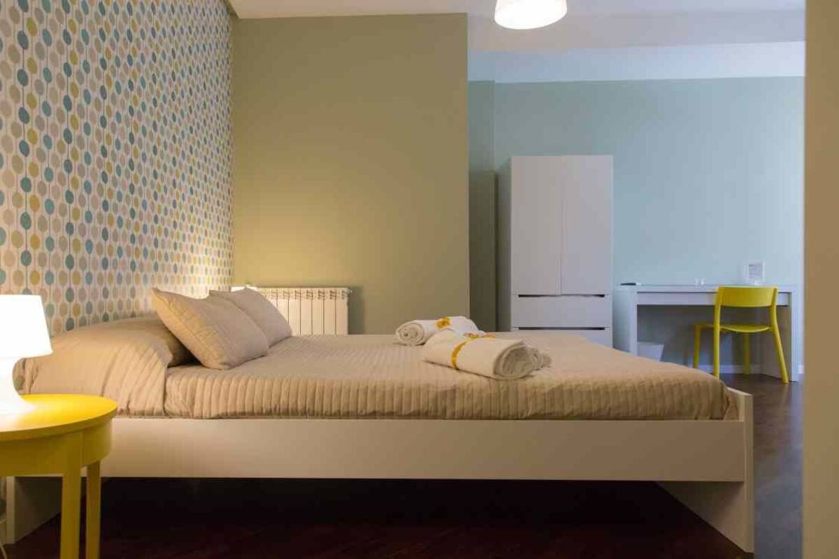 A bedroom with a bed, a bedside table, a lamp, a desk, and a chair.