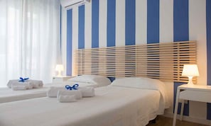 Una camera d'albergo con due letti, ognuno con copripiumino bianco e asciugamani blu. Sono presenti due comodini con lampade e un'ampia finestra con tende leggere.