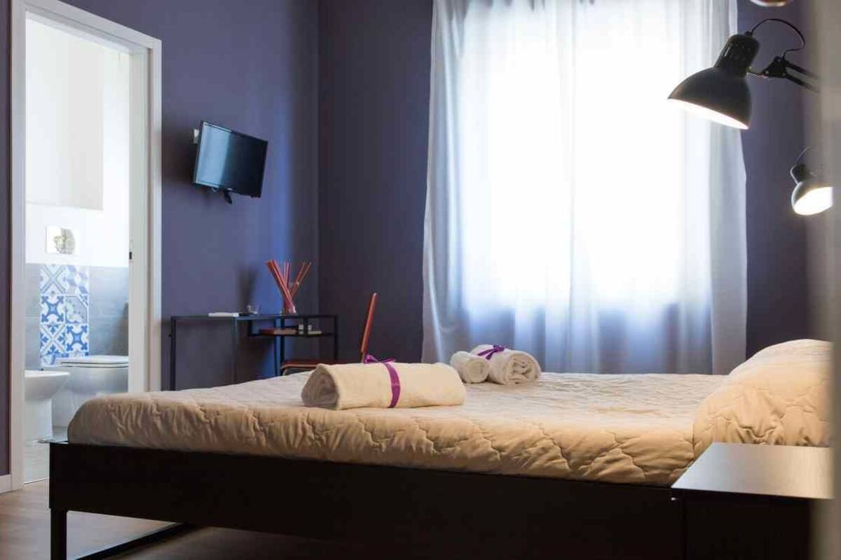 A bedroom with a bed, a television on the wall, a window with curtains, and a bedside lamp.