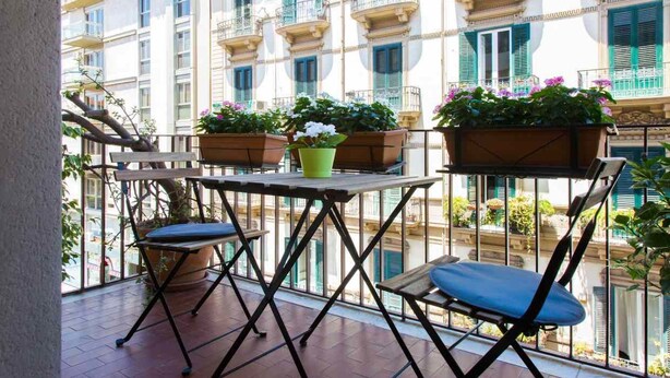 A balcony with a table and two chairs, potted plants, and a view of a building with balconies. - B&b Zammù (Palermo)