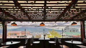 A restaurant with a view of a cityscape and mountains through large windows, featuring round tables and black chairs.