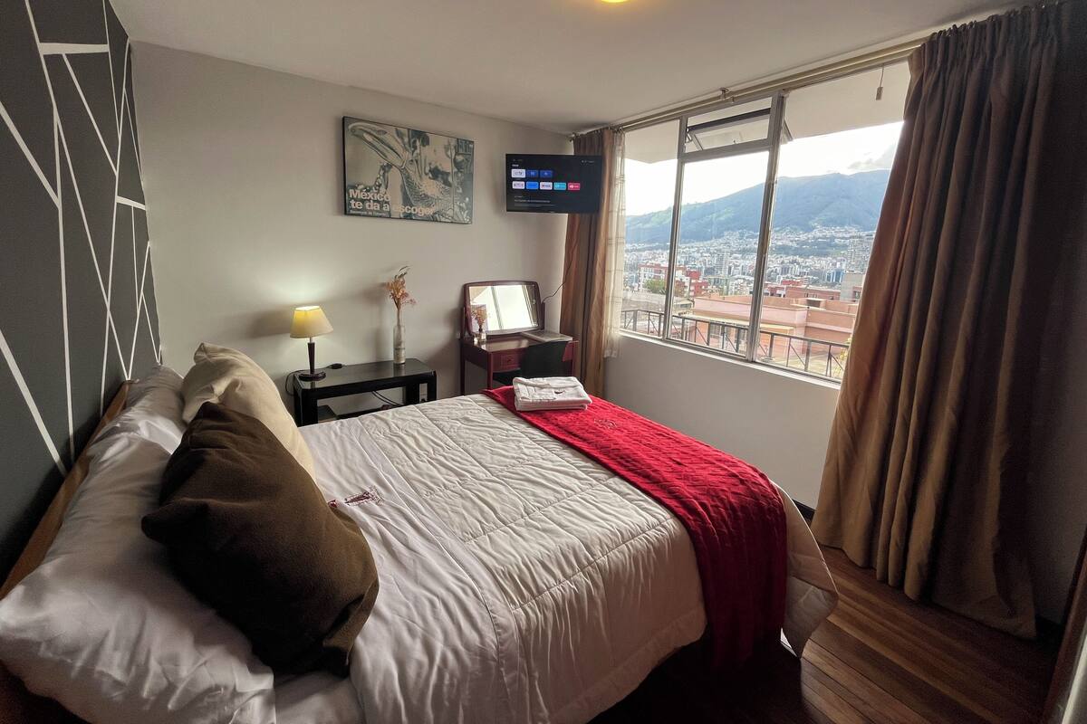 Habitación de hotel con cama, escritorio, silla, televisor y ventana con vista a la ciudad.