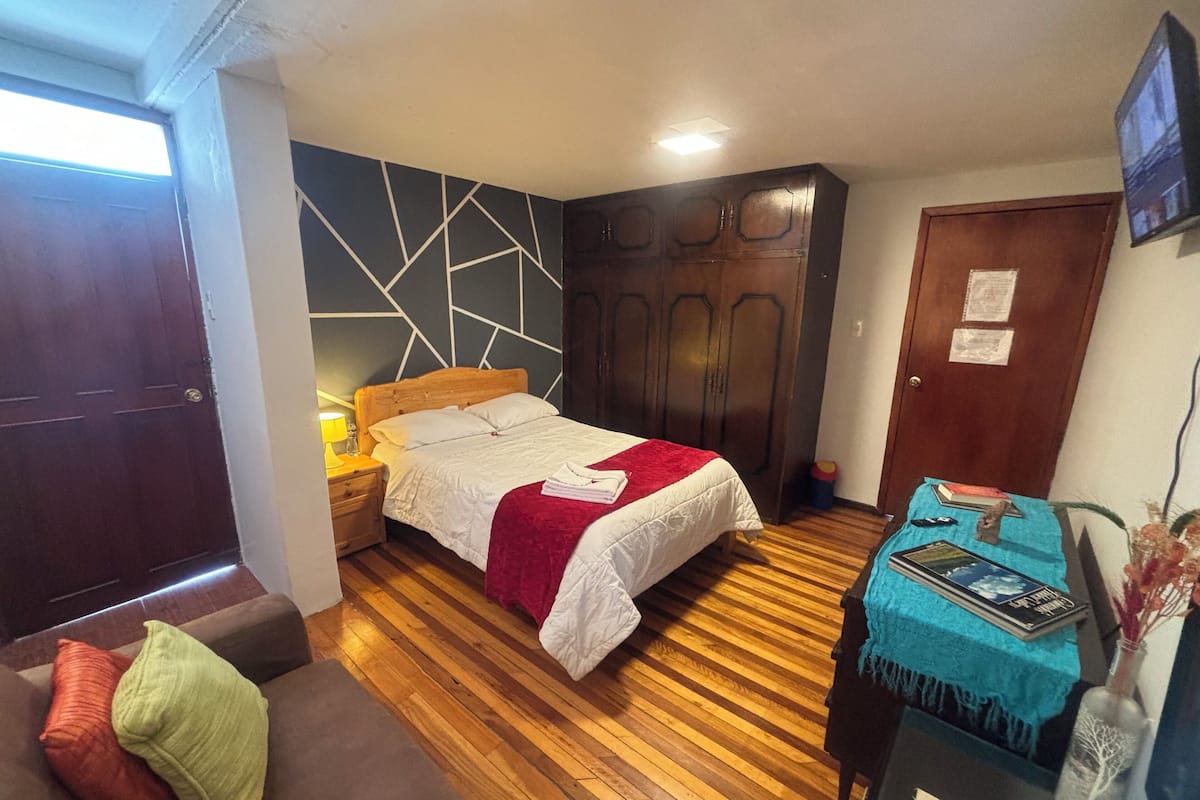 Un dormitorio con piso de madera, una cama con ropa blanca, un plaid rojo, una mesita de noche con una lámpara, un armario y una pequeña mesa con un florero y libros.