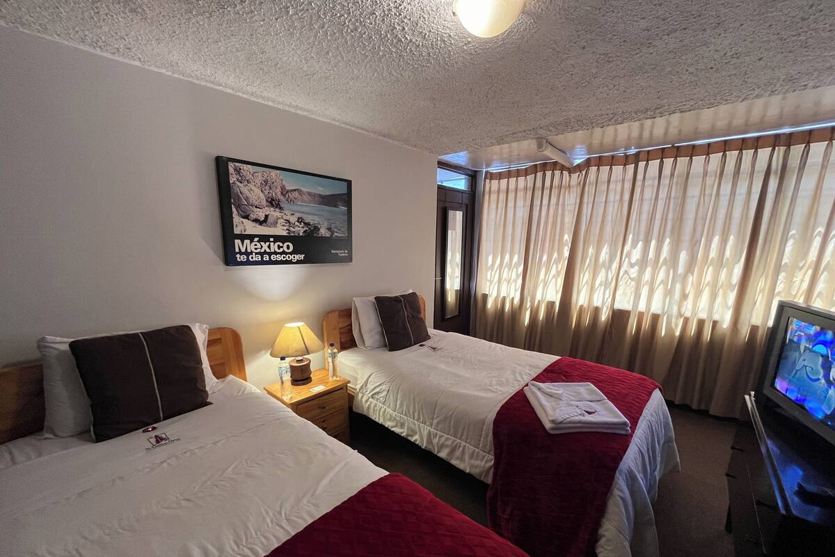 Habitación de hotel con dos camas, televisión, ventilador de techo y ventana con cortinas.