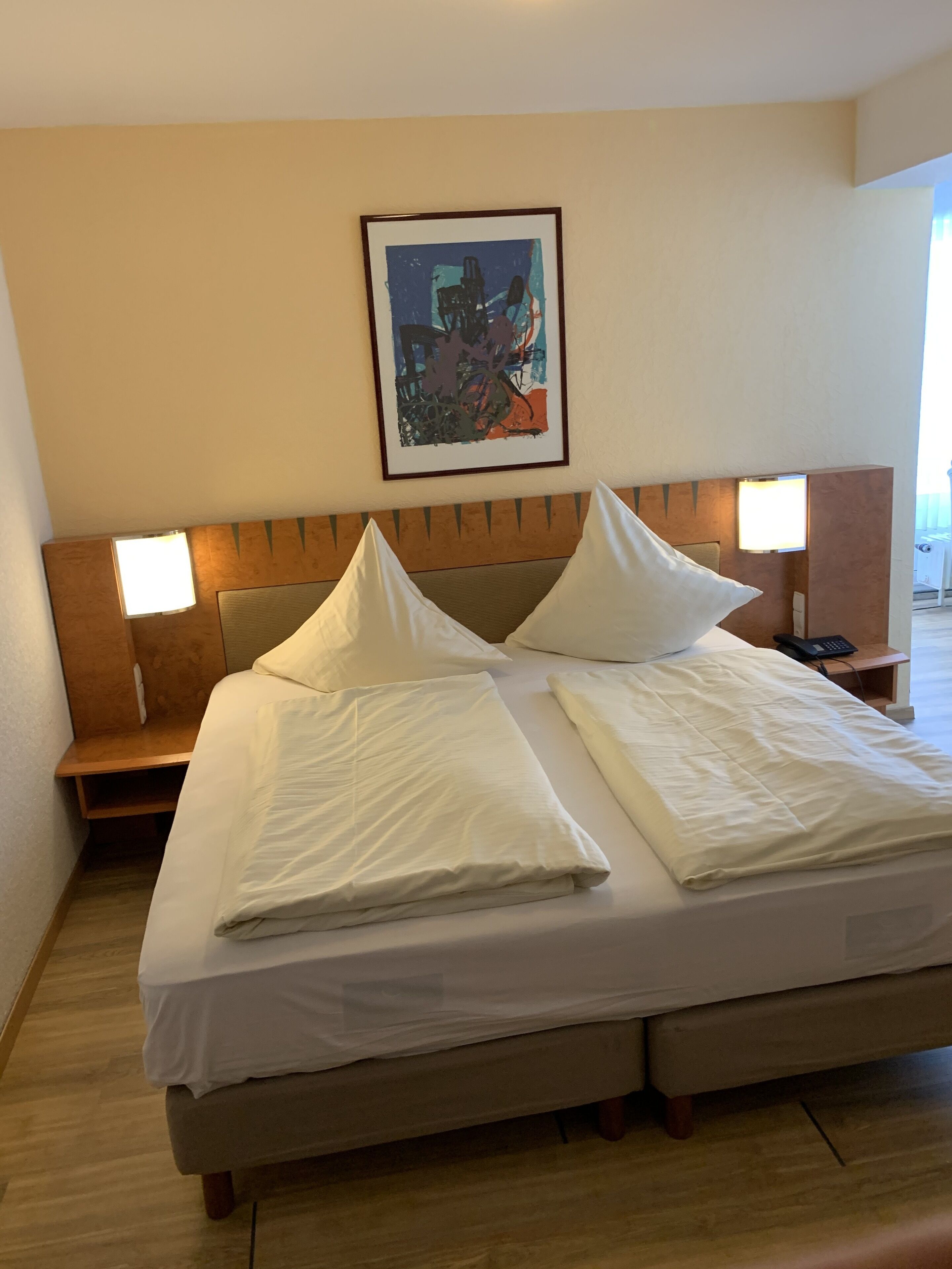 A neatly made bed with white linens, two bedside tables with lamps, and a framed abstract painting on the wall.