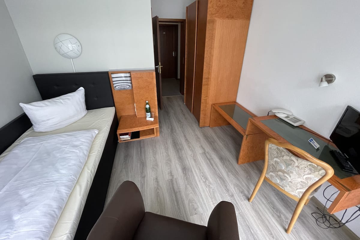 Una habitación de hotel con cama, escritorio, silla, armario y ventilador.