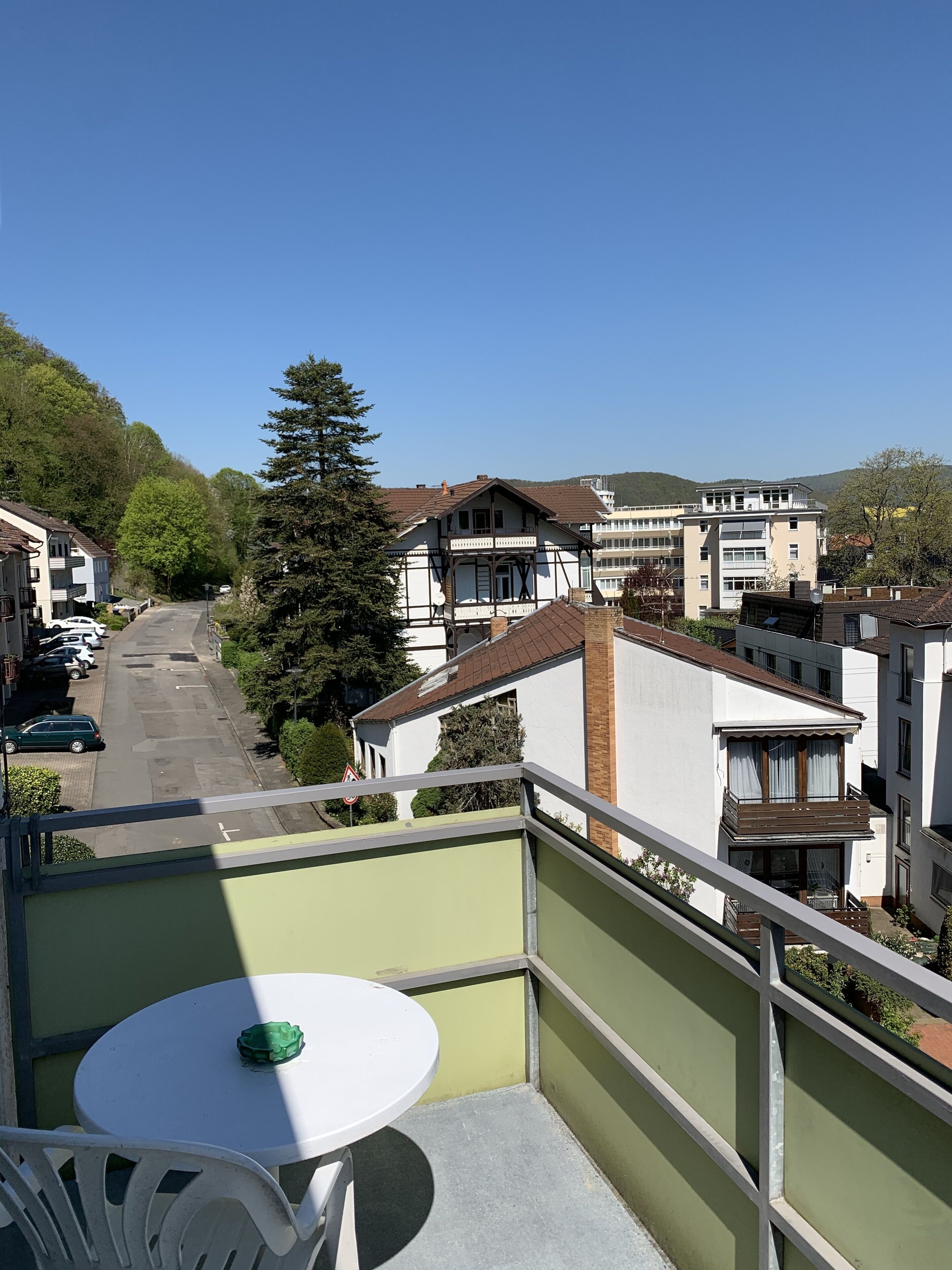Un balcón con mesa y sillas, con vista a una calle con coches aparcados y edificios.