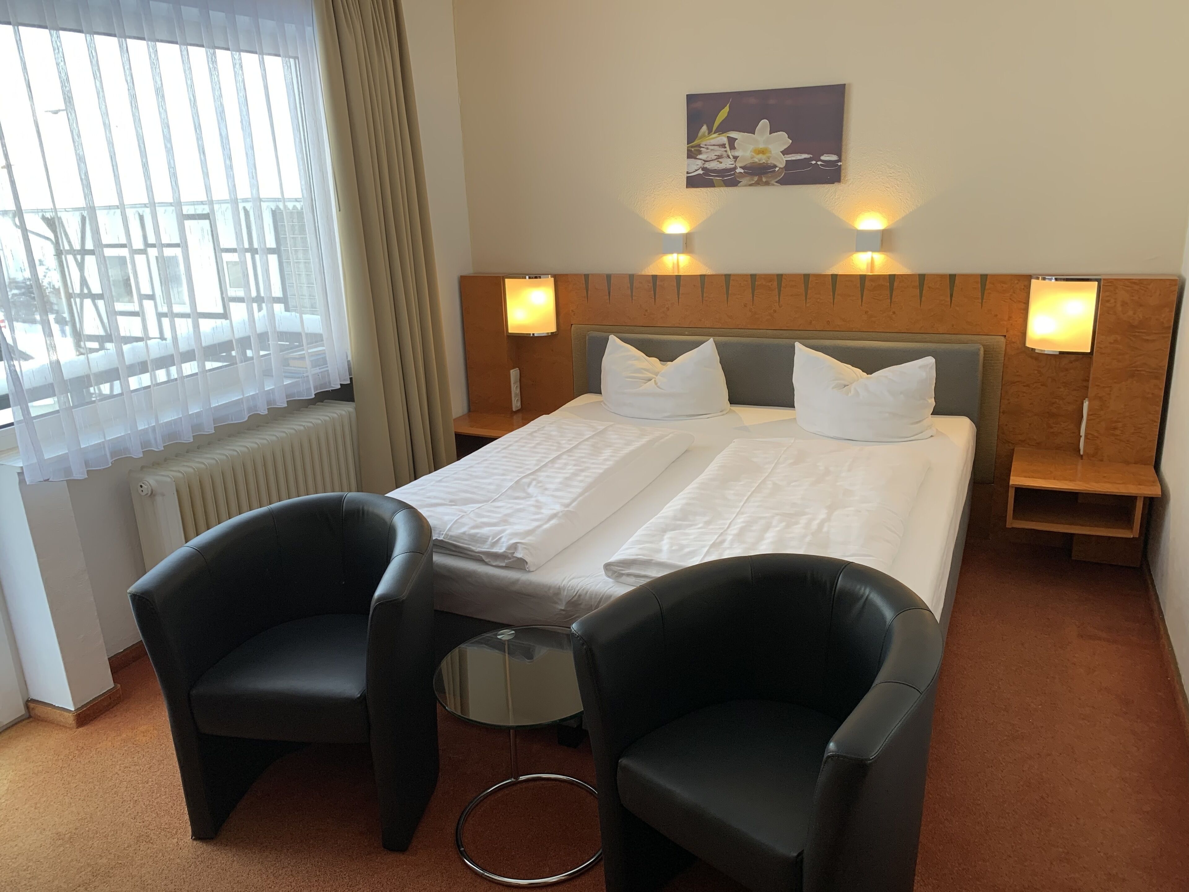 A hotel room with a bed, two armchairs, a small table, a radiator, a window with curtains, and wall-mounted lamps.