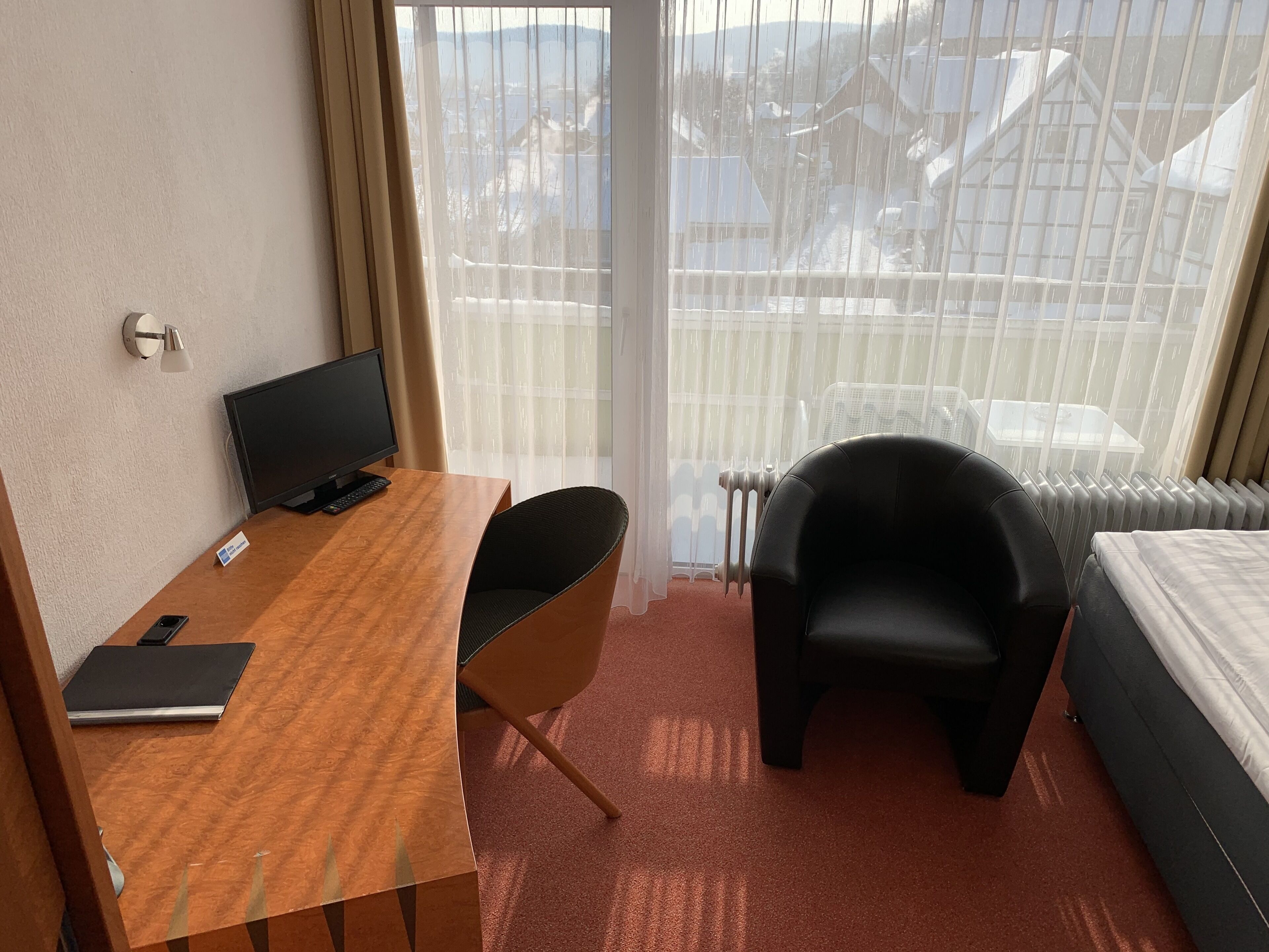 A hotel room with a wooden desk, a black chair, a bed, and a view of a snowy landscape through sheer curtains.