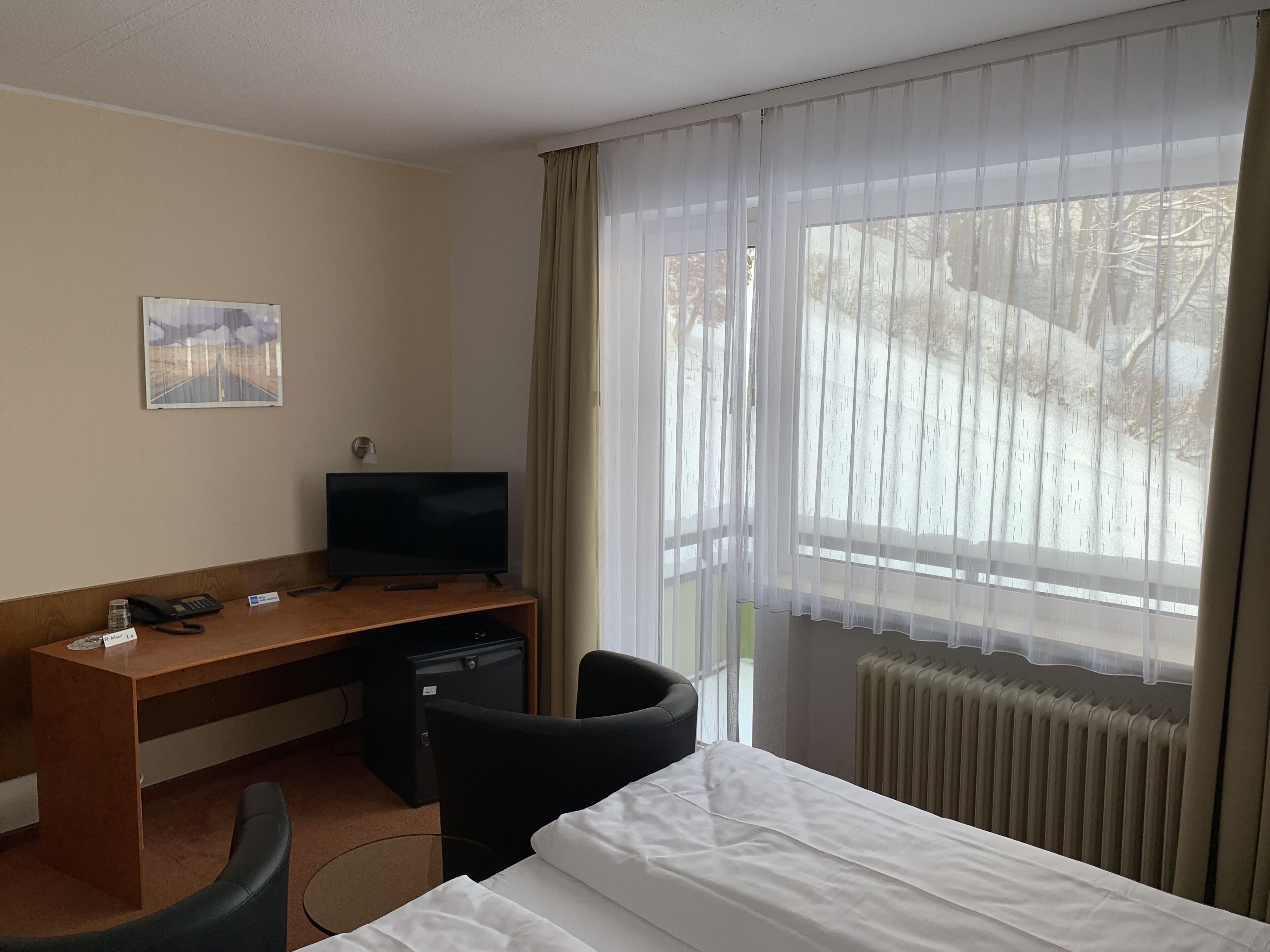 Ett hotellrum med en säng, ett skrivbord med en tv, en stol, ett litet skåp och ett fönster med gardiner.