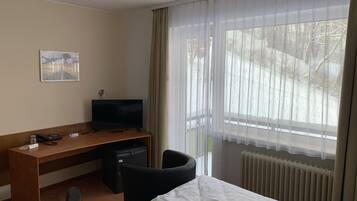 Ett hotellrum med en säng, ett skrivbord med en tv, en stol, ett litet skåp och ett fönster med gardiner.