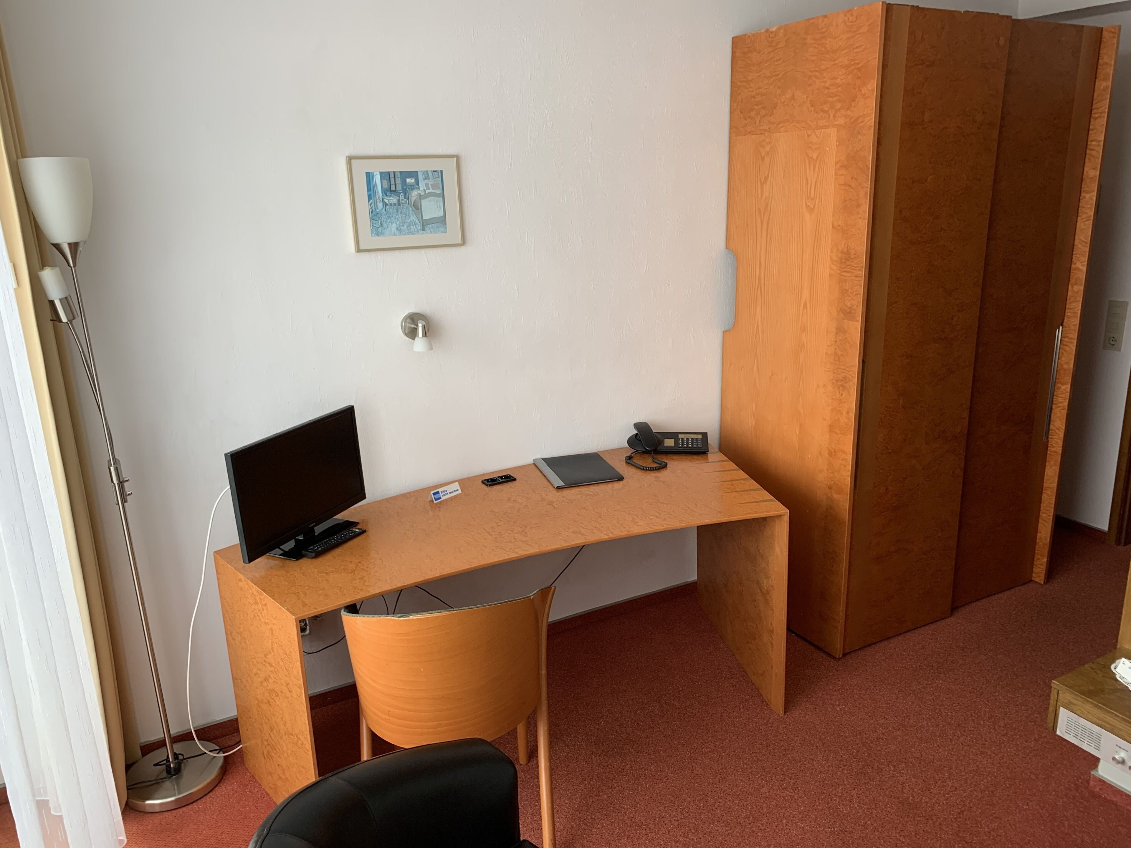 A room with a wooden desk, a black chair, a lamp, a phone, and a framed picture on the wall.