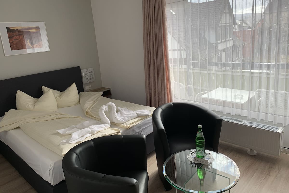 Habitación de hotel con cama, una silla negra, una mesita de vidrio con una botella y ventana con vista a edificios.