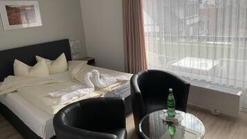 A hotel room with a bed, a black chair, a glass table with a bottle, and a window with a view of buildings.