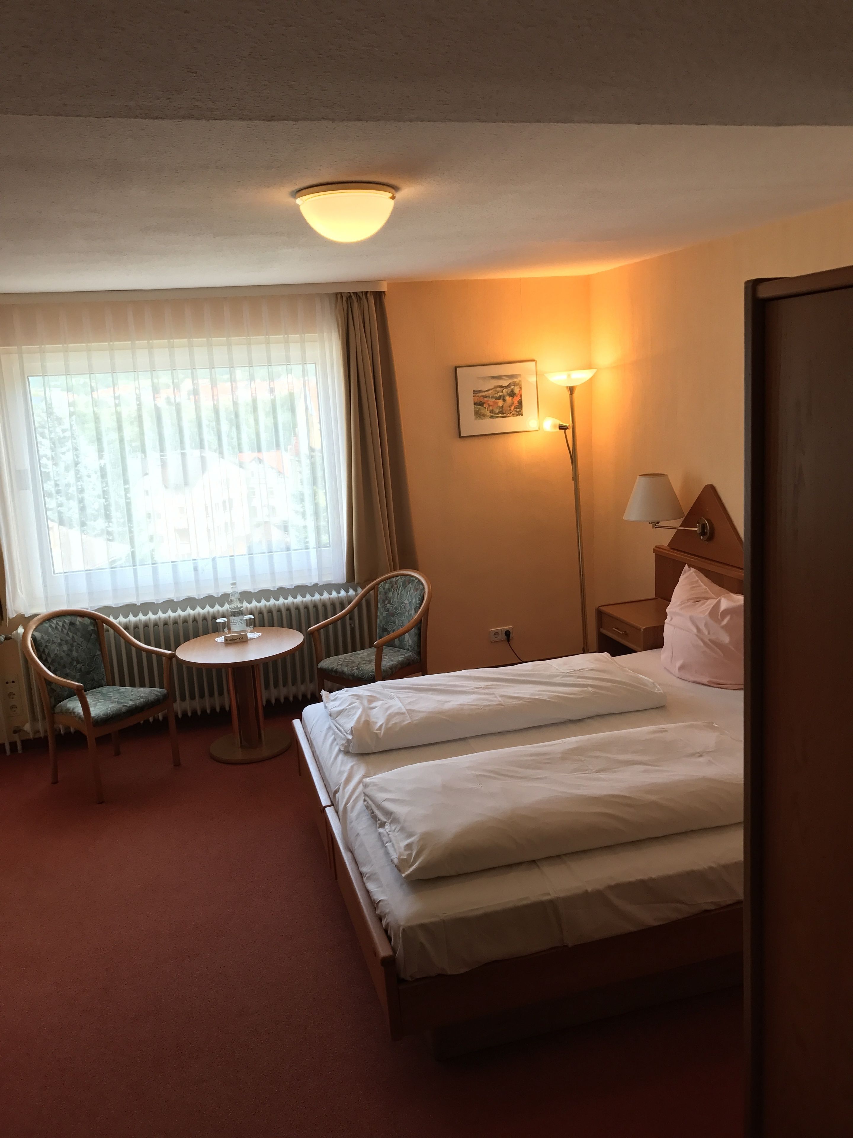 A hotel room with a bed, two chairs, a small table, a lamp, and a window with curtains.