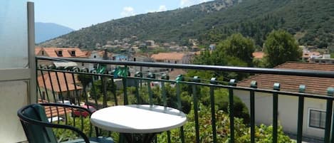 A balcony with two chairs and a table, overlooking a town with red-roofed buildings and a hill in the background.