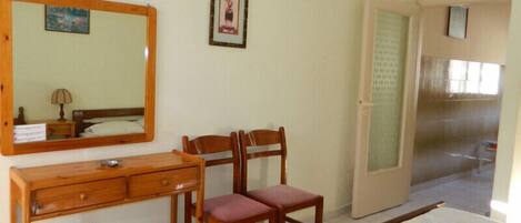A room with a wooden dresser, two wooden chairs, a bed, a mirror, and a door leading to another room.