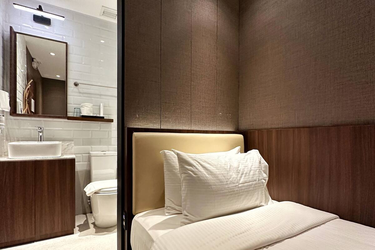 Una habitación de hotel moderna con una cama grande, un cabecero de madera, un baño con lavamanos y espejo, y una alfombra verde.