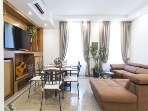 Una sala de estar con un sofá marrón, una mesa de comedor con sillas, un televisor sobre una estantería de madera y una guitarra.