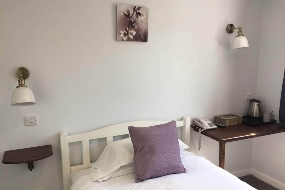 A neatly made bed with white linens and a purple pillow, a bedside table with a phone and a box labeled "TEA", and a wall-mounted lamp.