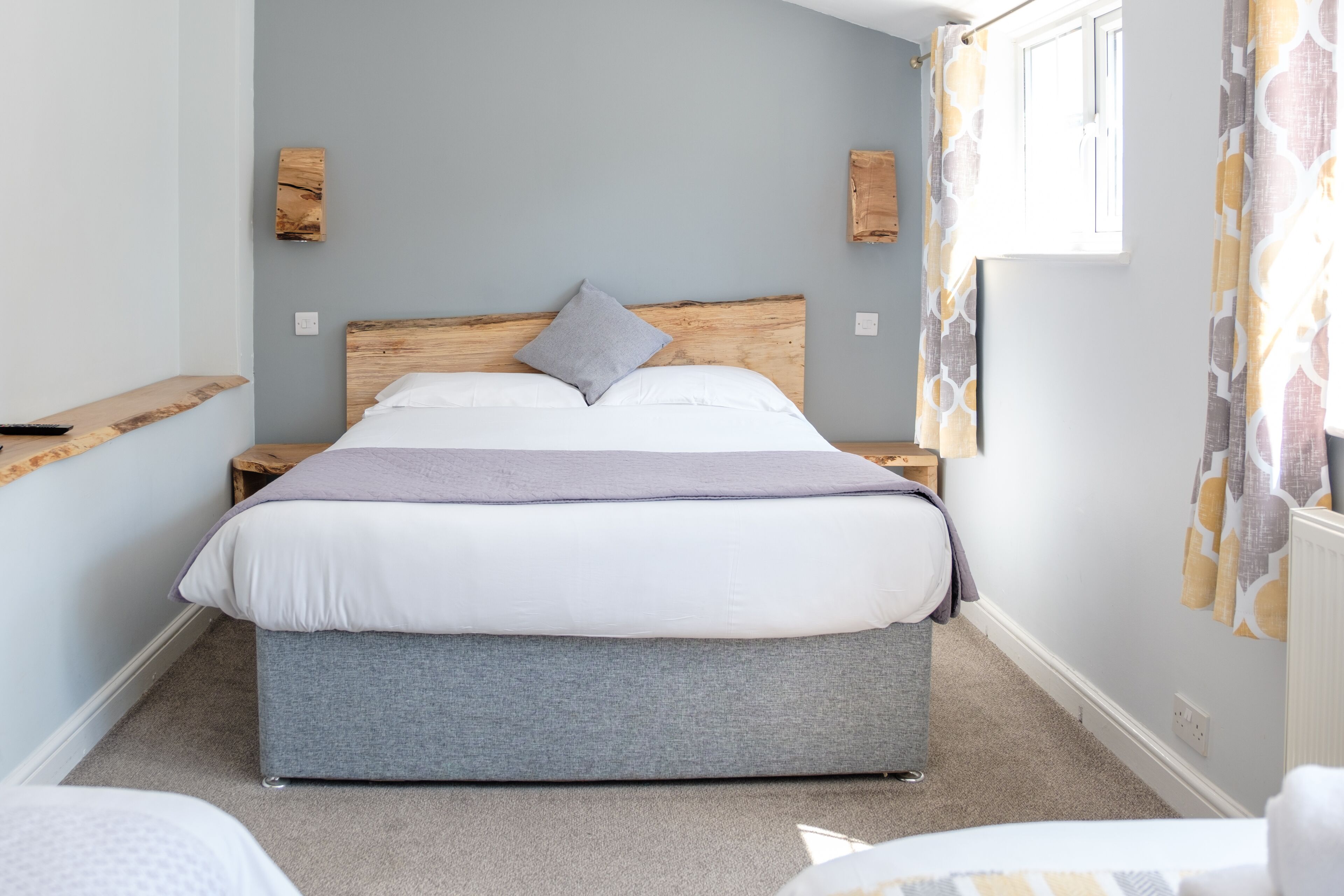 A bedroom with a large bed, a wooden headboard, a grey bench, and a window with curtains.