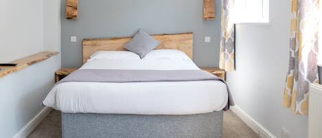 A bedroom with a large bed, a wooden headboard, a grey bench, and a window with curtains.