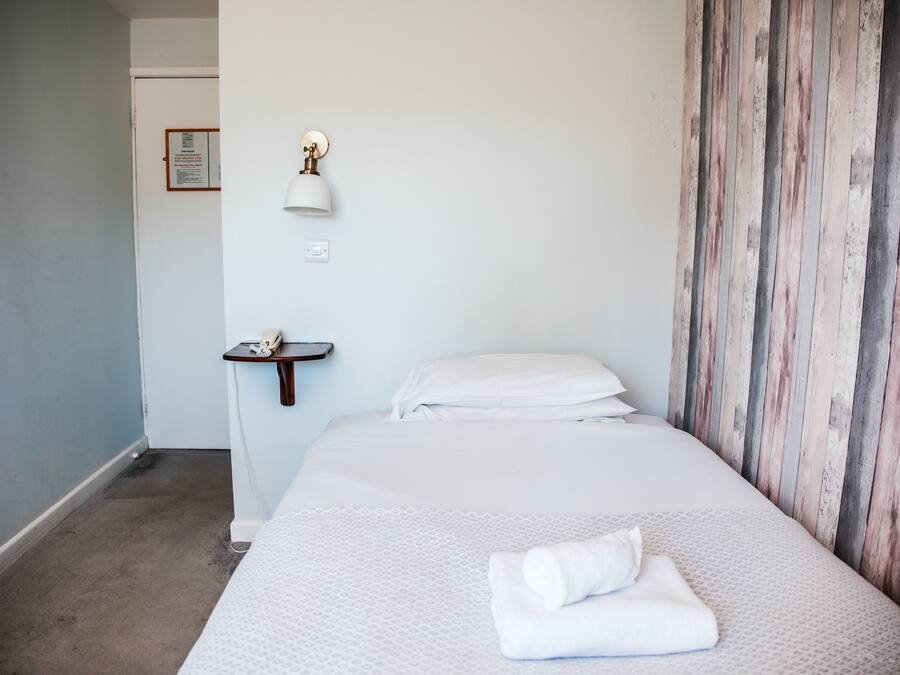 A hotel room with a bed, a small bedside table, a wall-mounted lamp, and a wooden panel wall.