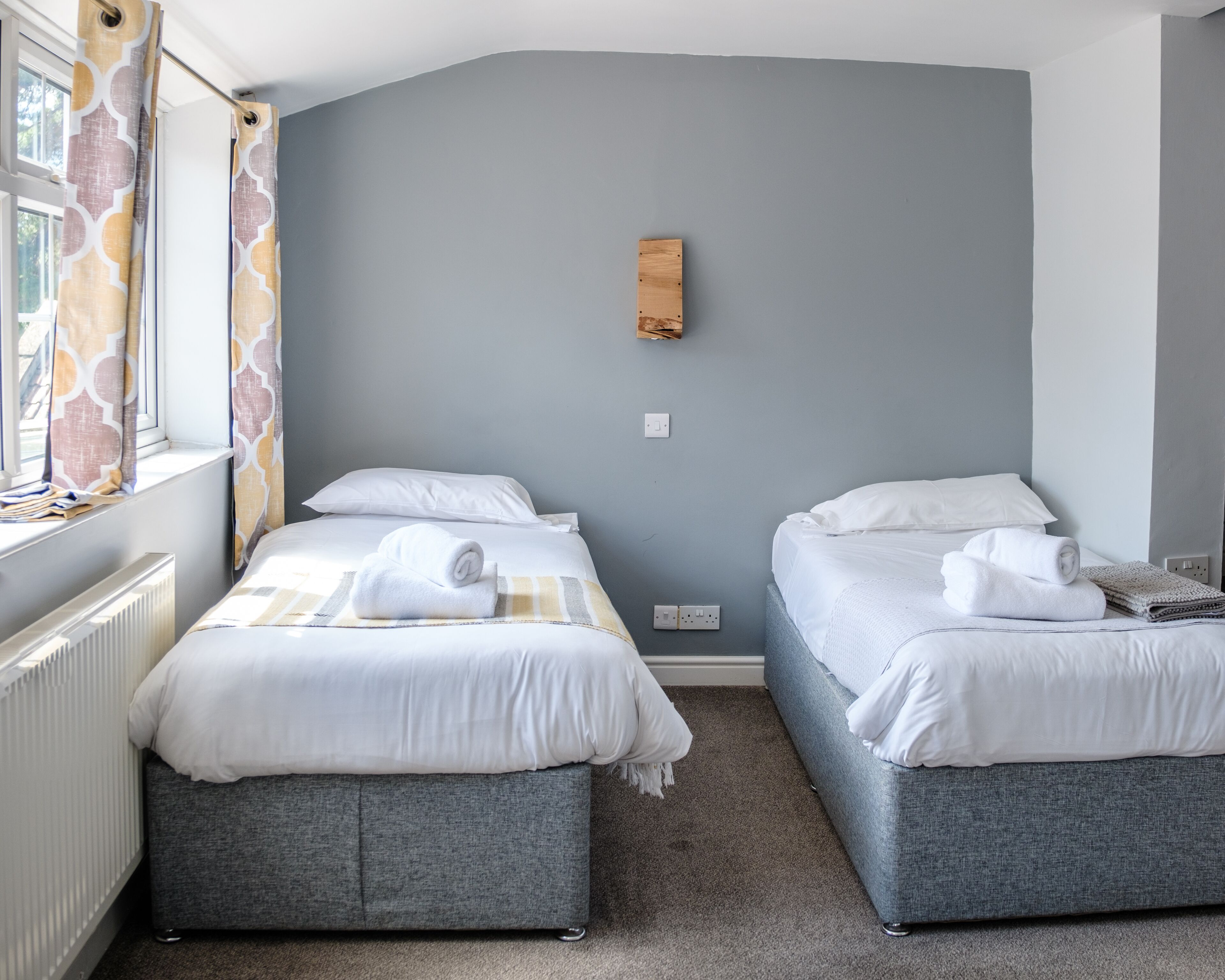A hotel room with two beds, grey upholstered headboards, white linens, and a window with patterned curtains.