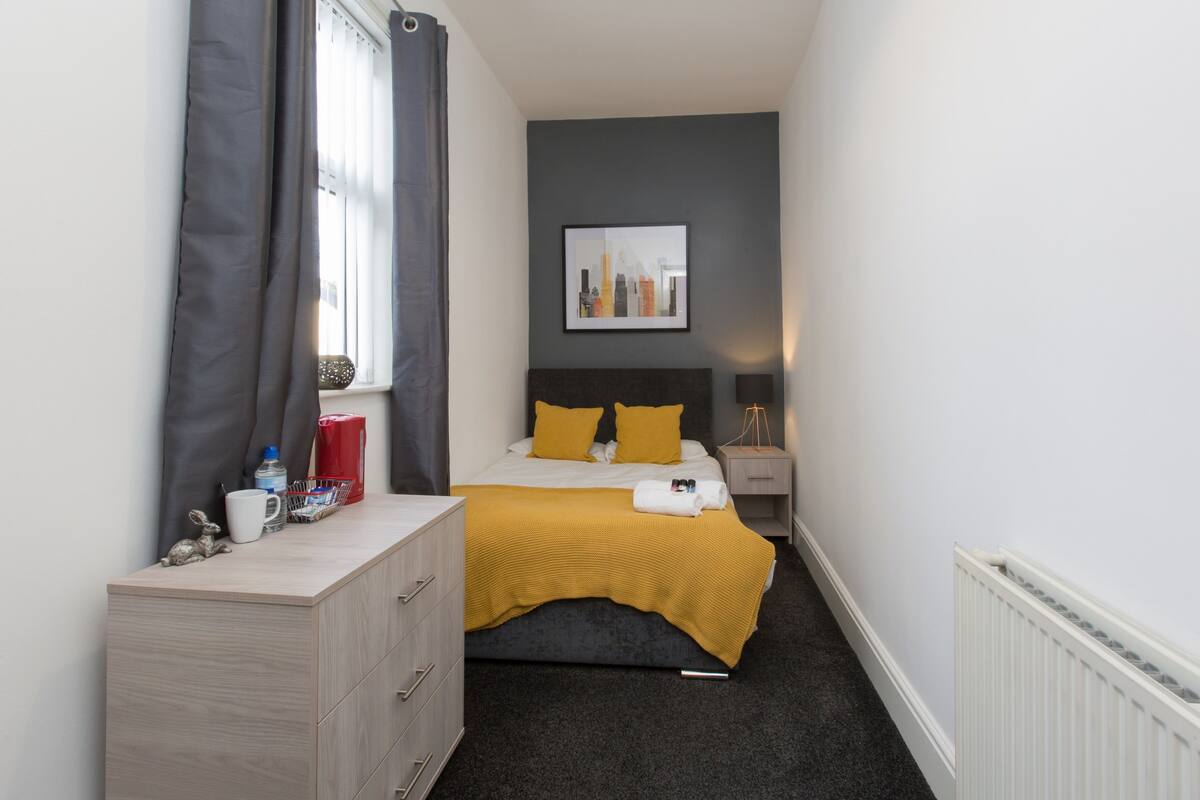 Une chambre d’hôtel étroite, avec un lit, une commode, une fenêtre avec des rideaux, un tableau encadré au mur et un radiateur.
